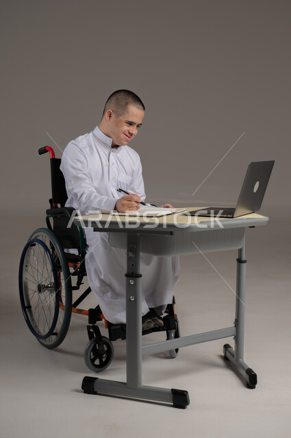 مفهوم حل الواجبات المدرسية والتعلم بواسطة التقنيات الحديثة, ذوي الاحتياجات الخاصة والهمم, بورتريه لشاب عربي خليجي سعودي يرتدي الثوب ويجلس على كرسي متحرك الدراسة بوسائل حديثة, خلفية رمادي