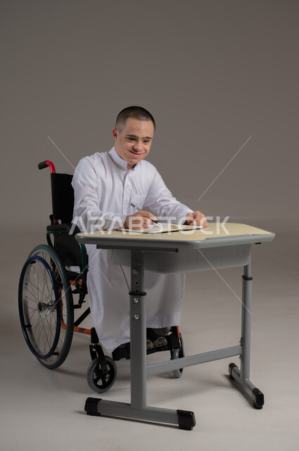 مفهوم حل الواجبات المدرسية والتعلم بواسطة التقنيات الحديثة, ذوي الاحتياجات الخاصة والهمم, بورتريه لشاب عربي خليجي سعودي يرتدي الثوب ويجلس على كرسي متحرك الدراسة بوسائل حديثة, خلفية رمادي
