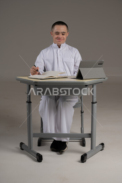 ذوي الاحتياجات الخاصة والهمم, مفهوم حل الواجبات المدرسية والتعلم بواسطة التقنيات الحديثة, بورتريه لشاب عربي خليجي سعودي يرتدي الثوب ويجلس على كرسي متحرك الدراسة بوسائل حديثة, خلفية رمادي