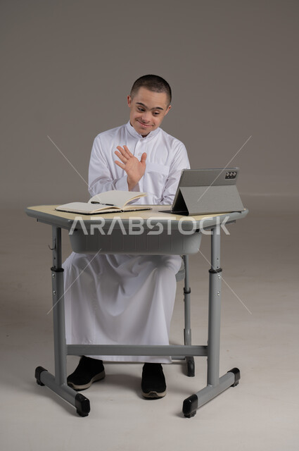 ذوي الاحتياجات الخاصة والهمم, مفهوم حل الواجبات المدرسية والتعلم بواسطة التقنيات الحديثة, بورتريه لشاب عربي خليجي سعودي يرتدي الثوب ويجلس على كرسي متحرك الدراسة بوسائل حديثة, خلفية رمادي