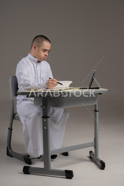 ذوي الاحتياجات الخاصة والهمم, مفهوم حل الواجبات المدرسية والتعلم بواسطة التقنيات الحديثة, بورتريه لشاب عربي خليجي سعودي يرتدي الثوب ويجلس على كرسي متحرك الدراسة بوسائل حديثة, خلفية رمادي