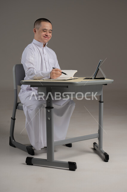 ذوي الاحتياجات الخاصة والهمم, مفهوم حل الواجبات المدرسية والتعلم بواسطة التقنيات الحديثة, بورتريه لشاب عربي خليجي سعودي يرتدي الثوب ويجلس على كرسي متحرك الدراسة بوسائل حديثة, خلفية رمادي