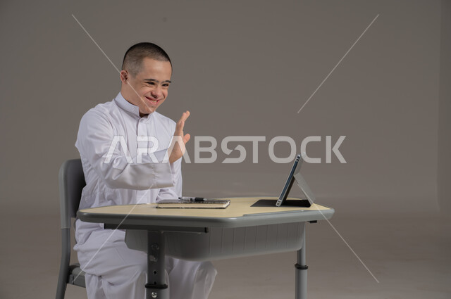 People with special needs and determination, the concept of doing schoolwork and learning using modern technologies, a portrait of a Saudi Gulf Arab young man wearing a thobe and sitting in a wheelchair, studying using modern means, gray background