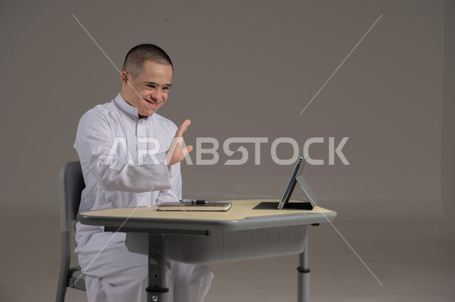 People with special needs and determination, the concept of doing schoolwork and learning using modern technologies, a portrait of a Saudi Gulf Arab young man wearing a thobe and sitting in a wheelchair, studying using modern means, gray background