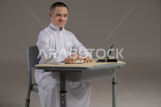 People with special needs and determination, the concept of doing schoolwork and learning using modern technologies, a portrait of a Saudi Gulf Arab young man wearing a thobe and sitting in a wheelchair, studying using modern means, gray background
