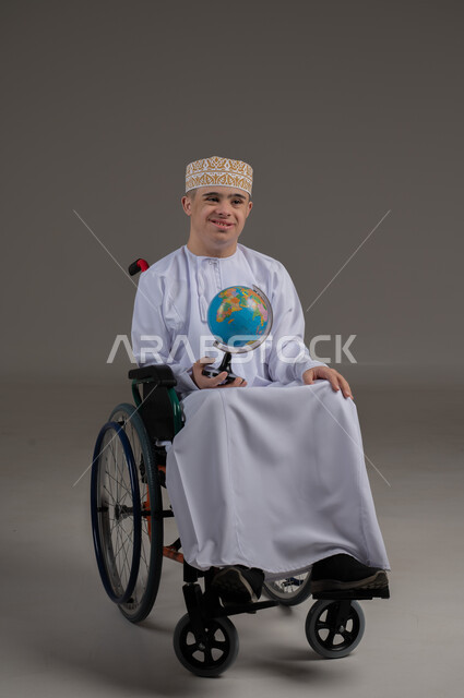 ذوي الاحتياجات الخاصة والهمم, النظر الى الكاميرا بتعابير السعادة والسرور, بورتريه لشاب عربي خليجي عماني يرتدي الكمة مع الدشداشة ويجلس على كرسي متحرك يحمل بيده كرة ارضية, خلفية رمادي