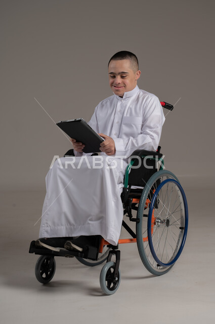 دعم واهتمام المجتمع العماني بأصحاب الاحتياجات الخاصه، استخدام الأجهزة التقنية والتكنولوجية الحديثة، بورتريه لولد عربي خليجي عماني مقعد مصاب بمتلازمة داون يرتدي الدشداشة يجلس على الكرسي المتحرك يحمل بيده جهازالتابلت، صورة لكامل الجسم، خلفية رمادية