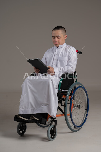 Using modern technical and technological devices, supporting and caring for people with special needs in Omani society, portrait of a disabled Gulf Arab Omani boy with Down syndrome wearing a dishdasha sitting in a wheelchair holding a tablet in his hand, full body image, gray background