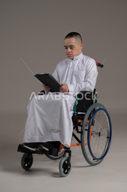 دعم واهتمام المجتمع العماني بأصحاب الاحتياجات الخاصه، استخدام الأجهزة التقنية والتكنولوجية الحديثة، بورتريه لولد عربي خليجي عماني مقعد مصاب بمتلازمة داون يرتدي الدشداشة يجلس على الكرسي المتحرك يحمل بيده جهازالتابلت، صورة لكامل الجسم، خلفية رمادية