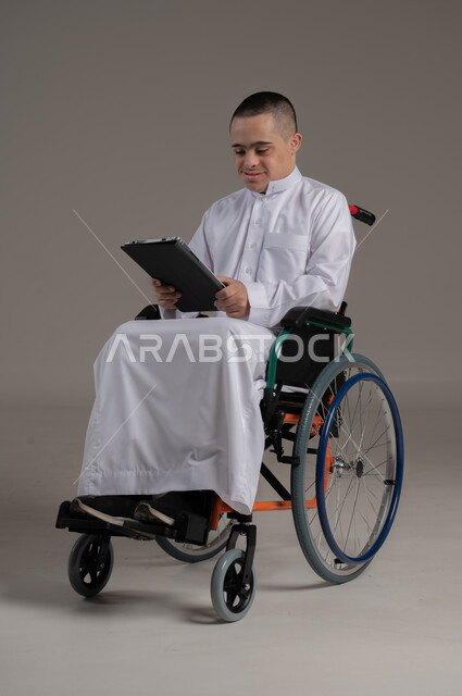 استخدام الأجهزة التقنية والتكنولوجية الحديثة، دعم واهتمام المجتمع العماني بأصحاب الاحتياجات الخاصه، بورتريه لولد عربي خليجي عماني مقعد مصاب بمتلازمة داون يرتدي الدشداشة يجلس على الكرسي المتحرك يحمل بيده جهازالتابلت، صورة لكامل الجسم، خلفية رمادية