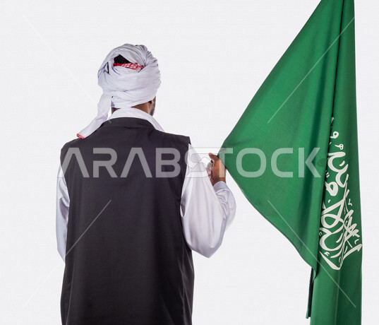 بورتريه بزاوية من الخلف لرجل عربي سوداني، يحمل بيده علم المملكة العربية السعودية، ذكرى اليوم الوطني السعودي، الاحتفال بالعيد الوطني السعودي، خلفية بيضاء لرجل سوداني مسلم يرتدي الزي السوداني مع العمه الملفحة