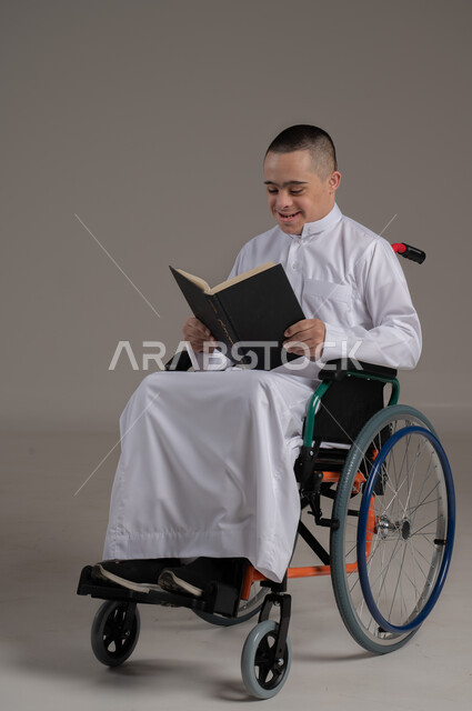 الأنشطة الترفيهية المفيدة لذوي الاحتياجات الخاصة, دعم المجتمع العماني لهوايات أصحاب الهمم, الاستمتاع بممارسة هواية المطالعة، بورتريه لشاب عربي خليجي عماني مقعد مصاب بمتلازمة داون يرتدي الدشداشة يجلس على كرسي متحرك يقوم بقراءة القصص, صورة لكامل الجسم، خلفية رمادية