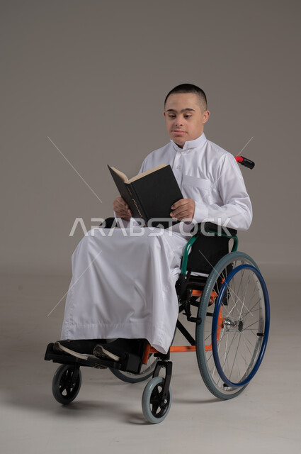 دعم المجتمع العماني لهوايات أصحاب الهمم, الاستمتاع بممارسة هواية المطالعة، الأنشطة الترفيهية المفيدة لذوي الاحتياجات الخاصة, بورتريه لشاب عربي خليجي عماني مقعد مصاب بمتلازمة داون يرتدي الدشداشة يجلس على كرسي متحرك يقوم بقراءة القصص, صورة لكامل الجسم، خلفية رمادية