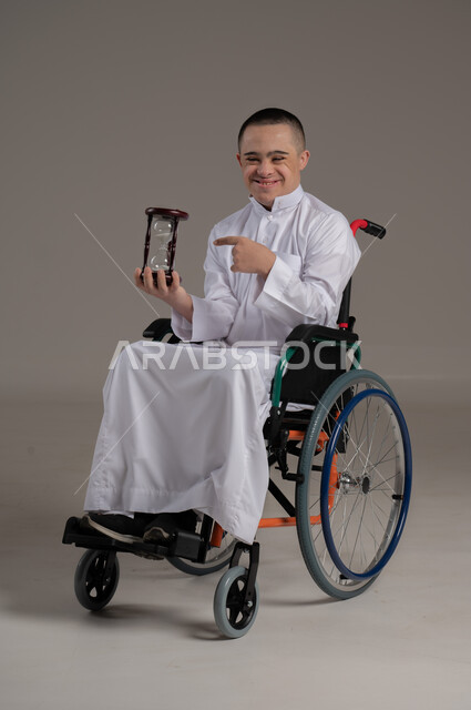 ادراك قيمة الوقت، القيام بمراقبة توقيت الرمل, مفهوم إدارة الأعمال وتنظيم الجدول الزمني، بورتريه لشاب عربي خليجي عماني مقعد مصاب بمتلازمة داون يرتدي الدشداشة يحمل بيده ساعة رملية زجاجية (البنكام) يشير اليها باصبعه، صورة الجسم بالكامل، خلفية رمادية