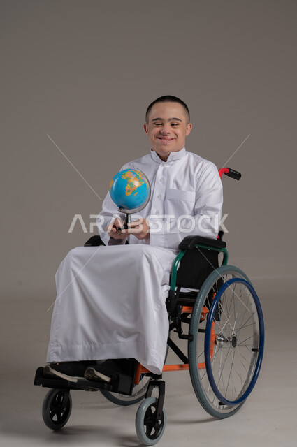 مفهوم البحث والاستكشاف حول العالم, القيام برحلة سياحية, بورتريه لشاب عربي خليجي عماني مقعد مصاب بمتلازمة داون يرتدي الدشداشة يجلس على كرسي متحرك يحمل بيده مجسم الكرة الارضية, صورة لكامل الجسم, خلفية رمادية