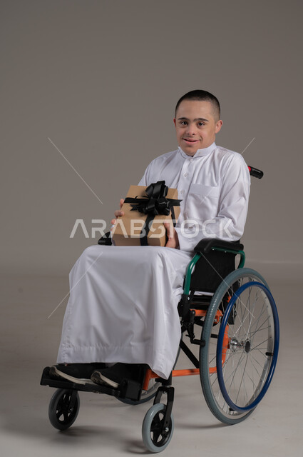 اهتمام ودعم المجتمع السعودي لذوي الهمم, مناسبات و أعياد سعيدة، بورتريه لشاب عربي خليجي عماني مقعد مصاب بمتلازمة داون يرتدي الدشداشة يجلس على كرسي متحرك يحمل بيده صندوق هدايا، شعور المعاقين بالحب و التقدير، صورة لكامل الجسم، خلفية رمادية