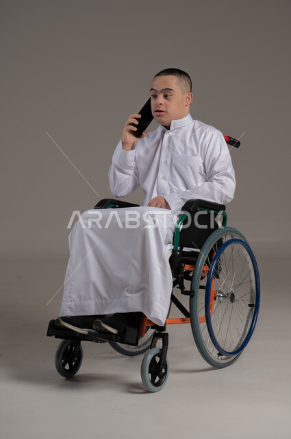 تعليم المعاقين على استخدام الاجهزة التقنية الحديثة، الدردشة والتواصل مع الاهل والأصدقاء، بورتريه لشاب عربي خليجي عماني مقعد مصاب بمتلازمة داون يرتدي الدشداشة يقوم بإجراء مكالمة هاتفية عبر الهاتف المحمول، ايماءات الفرح والسرور، صورة لكامل الجسم، خلفية رمادية