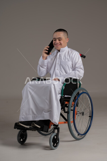 الدردشة والتواصل مع الاهل والأصدقاء، تعليم المعاقين على استخدام الاجهزة التقنية الحديثة، بورتريه لشاب عربي خليجي عماني مقعد مصاب بمتلازمة داون يرتدي الدشداشة يقوم بإجراء مكالمة هاتفية عبر الهاتف المحمول، ايماءات الفرح والسرور، صورة لكامل الجسم، خلفية رمادية