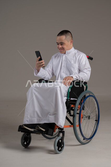 Teaching the disabled to use advanced technical devices, developing the capabilities of people with disabilities, portrait of a young Omani Gulf Arab man with a wheelchair, suffering from Down syndrome, wearing a dishdasha, using a mobile phone, expressions and gestures of integration and pleasure, full body image, gray background