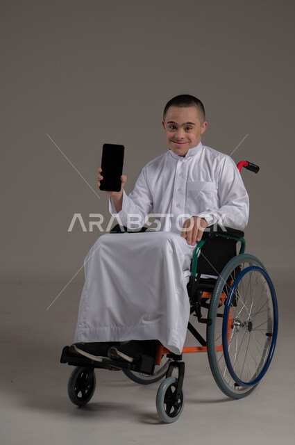 عرض شاشة سوداء فارغة، تعليم المعاقين على استخدام الأجهزة التقنية المتطورة، تنمية قدرات اصحاب الهمم، بورتريه لشاب عربي خليجي عماني مقعد مصاب بمتلازمة داون يرتدي الدشداشة يحمل الهاتف المحمول، تعابير وايماءات البهجة والسرور، صورة لكامل الجسم، خلفية رمادية