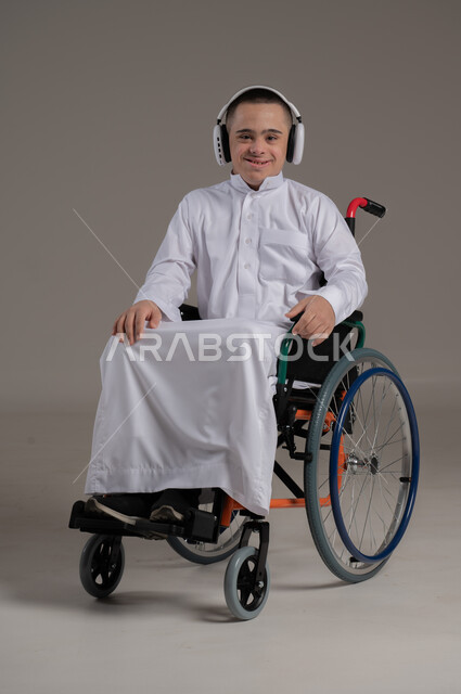 ايماءات السرور والسعادة، بورتريه لولد عربي خليجي عماني مقعد مصاب بمتلازمة داون يرتدي الدشداشة والسماعات، الاستماع الى الموسيقا والاغاني، مفهوم الترفيه والتسلية، نشاطات ترفيهية لذوي الهمم، صورة لكامل الجسم، خلفية رمادية