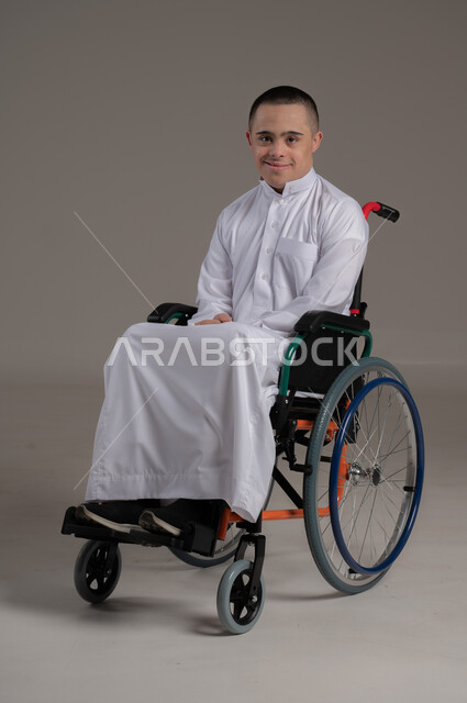 النظر إلى الكاميرا بايماءات الثقة بالنفس، مفهوم الاناقة والاهتمام بالمظهر الخارجي، اهتمام المجتمع العماني بذوي الهمم، بورتريه لشاب عربي خليجي عماني مقعد مصاب بمتلازمة داون يرتدي الدشداشة يجلس على كرسي متحرك، صورة لكامل الجسم، خلفية رمادية