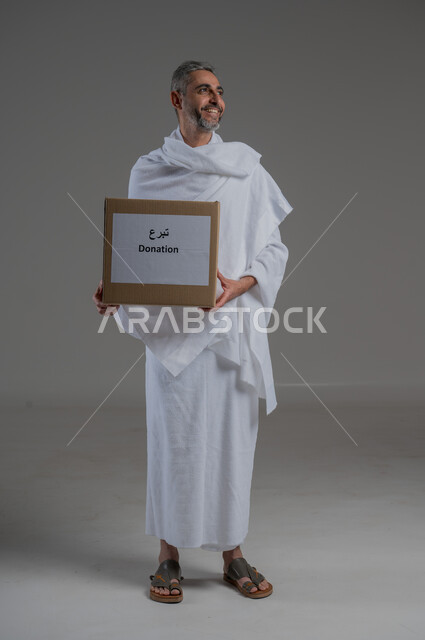 الأعمال الخيرية الإسلاميه، المساعدات والتبرعات الإنسانية، بورتريه لرجل عربي خليجي سعودي يرتدي زي الإحرام يحمل بيده صندوق تبرع، الدال على الخير كفاعله، الاحساس بالأخرين ومساعدتهم، أداء مناسك الحج والعمرة، صورة لكامل الجسم، خلفية رمادية