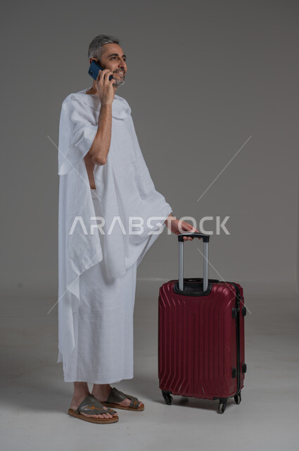 Heading and setting off to visit the Holy House of Allah, portrait of a Saudi Arabian Gulf man wearing Ihram clothes standing next to a red suitcase making a phone call, communicating with family and friends, performing Hajj and Umrah, using modern technology, full body image, gray background
