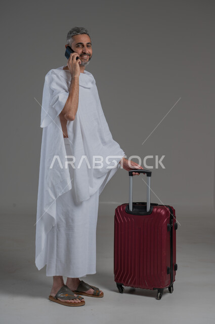 التواصل مع الاهل والاصدقاء، التوجه والانطلاق لزيارة بيت الله الحرام, بورتريه  لرجل عربي خليجي سعودي يرتدي ملابس الاحرام يقف بجانب حقيبة سفر حمراء يقوم باجراء مكالمة هاتفية، أداء فريضة الحج و العمرة، استخدام التكنولوجيا الحديثة، صورة لكامل الجسم، خلفية رمادية