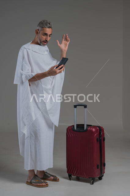 ايماءات الغضب والانفعال، التوجه والانطلاق لزيارة بيت الله الحرام, بورتريه  لرجل عربي خليجي سعودي يرتدي ملابس الاحرام يقف بجانب حقيبة سفر حمراء يحمل بيده الهاتف المحمول، أداء فريضة الحج و العمرة، استخدام التكنولوجيا الحديثة، صورة لكامل الجسم، خلفية رمادية