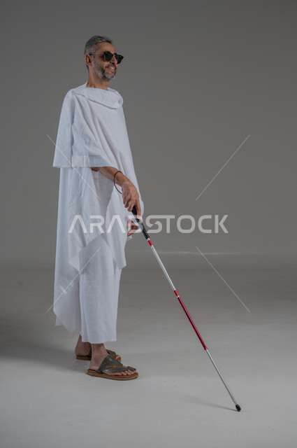 أداء فريضة الحج و العمرة، ايماءات السرور والسعادة، ضعف القدرة البصرية، بورتريه  لرجل عربي خليجي سعودي كفيف مصاب بالعمى يرتدي ملابس الاحرام والنظارة الشمسية يمسك بيده عصا، صورة لكامل الجسم، خلفية رمادية