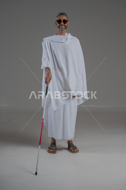 ضعف القدرة البصرية، ايماءات السرور والسعادة، بورتريه  لرجل عربي خليجي سعودي كفيف مصاب بالعمى يرتدي ملابس الاحرام والنظارة الشمسية يمسك بيده عصا، أداء فريضة الحج و العمرة، صورة لكامل الجسم، خلفية رمادية