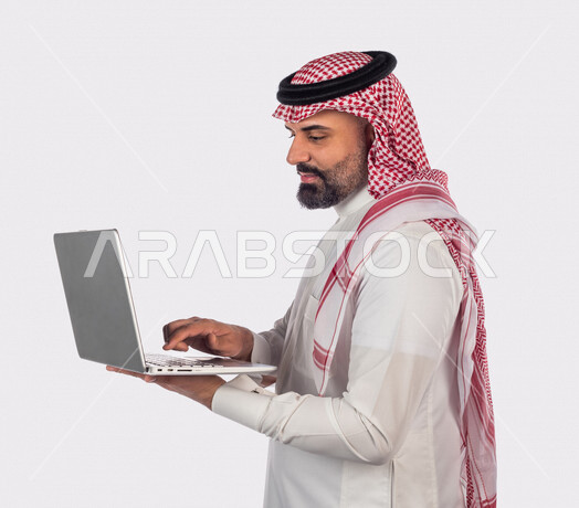 بورتريه لرجل سعودي خليجي بالزي السعودي التقليدي ، استخدام جهاز الحاسوب المحمول لإنجاز المهام والمشاريع ، خلفية بيضاء ، جلسة عمل، سيد أعمال، إنجاز المشاريع والمهام، العمل عن بعد ، مهن وأعمال تجارية ، التعليم الإلكتروني