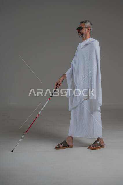 ضعف القدرة البصرية، ايماءات السرور والسعادة، بورتريه  لرجل عربي خليجي سعودي كفيف مصاب بالعمى يرتدي ملابس الاحرام والنظارة الشمسية يمسك بيده عصا، أداء فريضة الحج و العمرة، صورة لكامل الجسم، خلفية رمادية