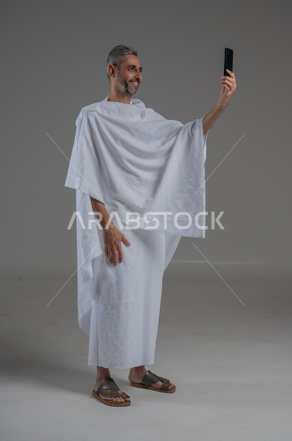 Using modern technical devices, making a video call, taking souvenir photos, communicating with family and friends, portrait of a Saudi Arabian Gulf man wearing Ihram clothes holding a mobile phone, performing Hajj and Umrah, full body photo, gray background