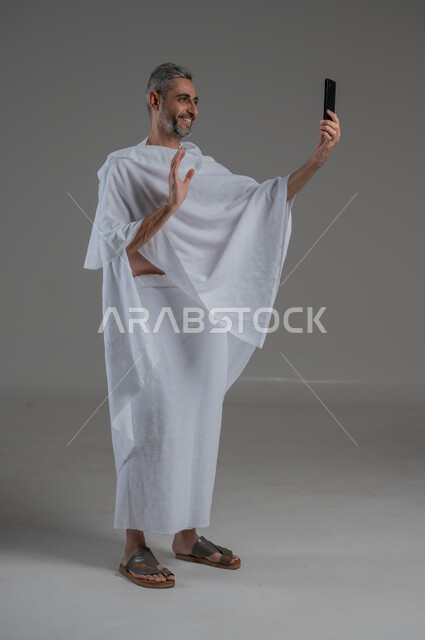 اجراء مكالمة فيديو، التقاط الصور التذكارية، التواصل مع الاهل والاصدقاء، استخدام الاجهزة التقنية الحديثة، بورتريه  لرجل عربي خليجي سعودي يرتدي ملابس الاحرام يحمل بيده الهاتف المحمول، أداء فريضة الحج و العمرة، صورة لكامل الجسم، خلفية رمادية