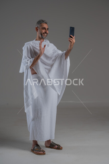 Taking souvenir photos, making a video call, communicating with family and friends, using modern technical devices, portrait of a Saudi Arabian Gulf man wearing Ihram clothes holding a mobile phone, performing Hajj and Umrah, full body photo, gray background