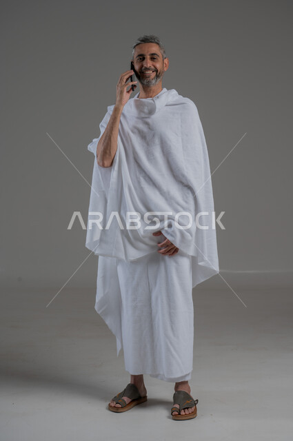 Communicating with family and friends, gestures of pleasure and happiness, making phone calls, using modern technical devices, portrait of a Saudi Arabian Gulf man wearing Ihram clothes holding a mobile phone, performing Hajj and Umrah, full body photo, gray background