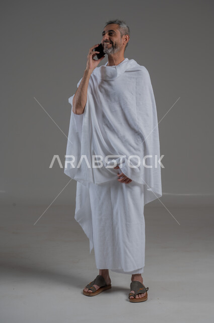 Making phone calls, gestures of pleasure and happiness, using modern technical devices, communicating with family and friends, portrait of a Saudi Arabian Gulf man wearing Ihram clothes holding a mobile phone, performing Hajj and Umrah, full body image, gray background