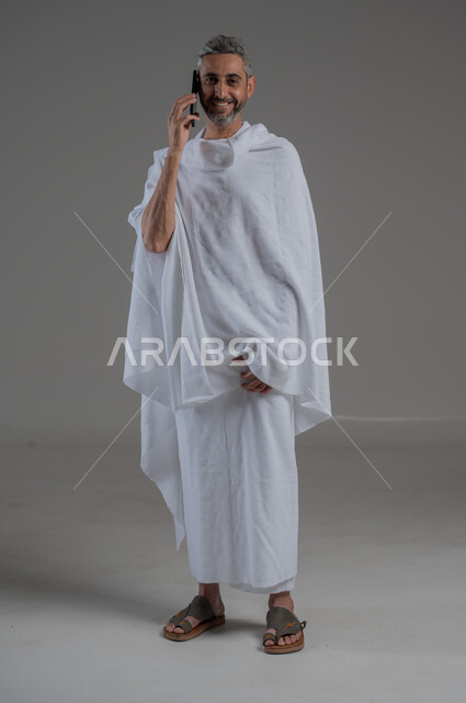 اجراء مكالمات هاتفية، ايماءات السروروالسعادة، استخدام الاجهزة التقنية الحديثة، التواصل مع الاهل والاصدقاء، بورتريه  لرجل عربي خليجي سعودي يرتدي ملابس الاحرام يحمل بيده الهاتف المحمول، أداء فريضة الحج و العمرة، صورة لكامل الجسم، خلفية رمادية