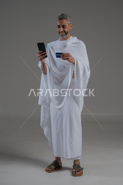 الحجز الالكتروني في حملات الحج والعمرة, الشراء والدفع عن طريق بطاقة الصراف الإئتمانية, بورتريه لشاب عربي خليجي سعودي يرتدي زي الاحرام يحمل بيده الهاتف الجوال وكرت البنك المصرفي, حجوزات الاونلاين السريعة، صورة لكامل الجسم، خلفية رمادية