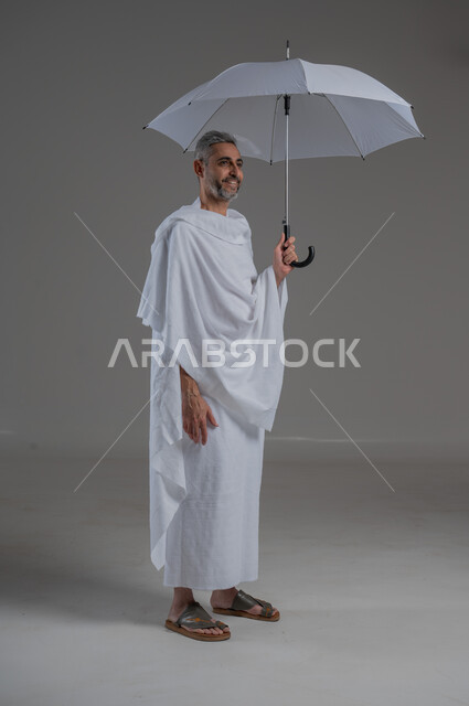 ايماءات البهجة والسرور، زيارة بيت الله الحرام في مكة المكرمة، بورتريه لرجل عربي خليجي سعودي يرتدي ملابس الاحرام يحمل بيده مظلة للحماية من أشعة الشمس، الركن الخامس من أركان الاسلام، آداء مناسك الحج والعمرة، صورة لكامل الجسم، خلفية رمادية