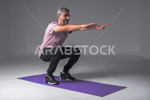 تقوية الأعصاب والعضلات، اتباع أسلوب حياة صحي، المحافظة على التوازن الجسدي، ايماءات القوة والنشاط، مفهوم اللياقة والرشاقة، بورتريه لرجل عربي خليجي سعودي يرتدي الزي الرياضي يقوم بممارسة التمرينات الرياضية، خلفية رمادية.