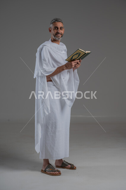 تلاوة المصحف الشريف، قراءة كتاب الله وحفظه وتجويده، مفهوم الاجتهاد في العبادة، الركن الخامس من أركان الاسلام، بورتريه من الجانب لرجل عربي خليجي سعودي يرتدي ملابس الاحرام يحمل بيده القرآن، آداء مناسك الحج والعمرة، صورة لكامل الجسم، خلفية رمادية