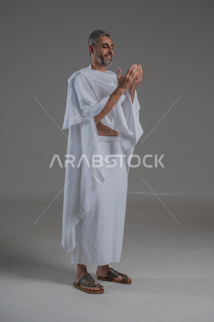 التضرع إلى الله تعالى، تعابير التوسل والاسترحام، طلب المساعدة من المولى عز وجل، مفهوم الاجتهاد في العبادة، ، بورتريه من الجانب لرجل عربي خليجي سعودي يرتدي ملابس الاحرام يرفع ييه بايماءات الدعاء، آداء مناسك الحج والعمرة، صورة لكامل الجسم، خلفية رمادية