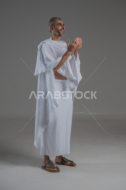تعابير التوسل والاسترحام، التضرع إلى الله تعالى، طلب المساعدة من المولى عز وجل، مفهوم الاجتهاد في العبادة، ، بورتريه من الجانب لرجل عربي خليجي سعودي يرتدي ملابس الاحرام يرفع ييه بايماءات الدعاء، آداء مناسك الحج والعمرة، صورة لكامل الجسم، خلفية رمادية