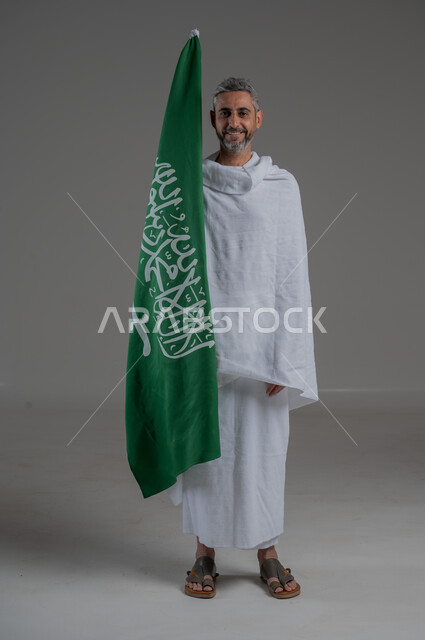 يوم العلم 11 مارس، اليوم الوطني السعودي 23 سبتمبر، الفخر و الاعتزاز بالانتماء للوطن، بورتريه لرجل عربي خليجي سعودي يرتدي ملابس الاحرام يحمل بيده راية التوحيد والسلام، اداء مناسك الحج والعمرة، صورة لكامل الجسم، خلفية رمادية