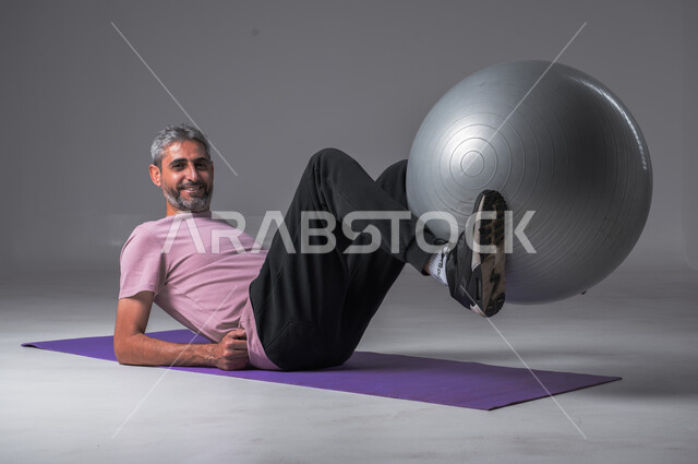 النظر الى الكاميرا بايماءات السعاده ، أسلوب حياة صحي ، تقوية عضلات الجسم ، نشاط بدني للمحافظة على الصحة و اللياقة الجسدية ، بورتريه لرجل عربي خليجي سعودي يقوم بممارسة التمارين الرياضية مع كرة بيلاتيس ، خلفيه رماديه