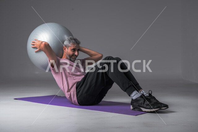 Healthy lifestyle, strengthening body muscles, physical activity to maintain health and physical fitness, portrait of a Saudi Arabian Gulf man doing exercises with a Pilates ball, gray background