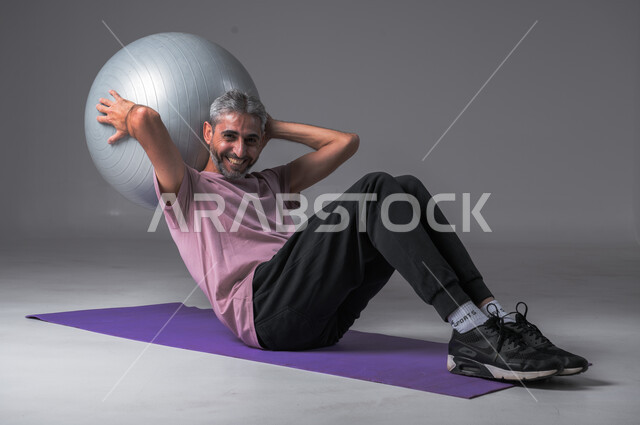 ايماءات وجه تدل على السعادة ، نشاط بدني للمحافظة على الصحة و اللياقة الجسدية ، أسلوب حياة صحي ، تقوية عضلات الجسم ، بورتريه لرجل عربي خليجي سعودي يقوم بممارسة التمارين الرياضية مع كرة بيلاتيس ، خلفيه رماديه